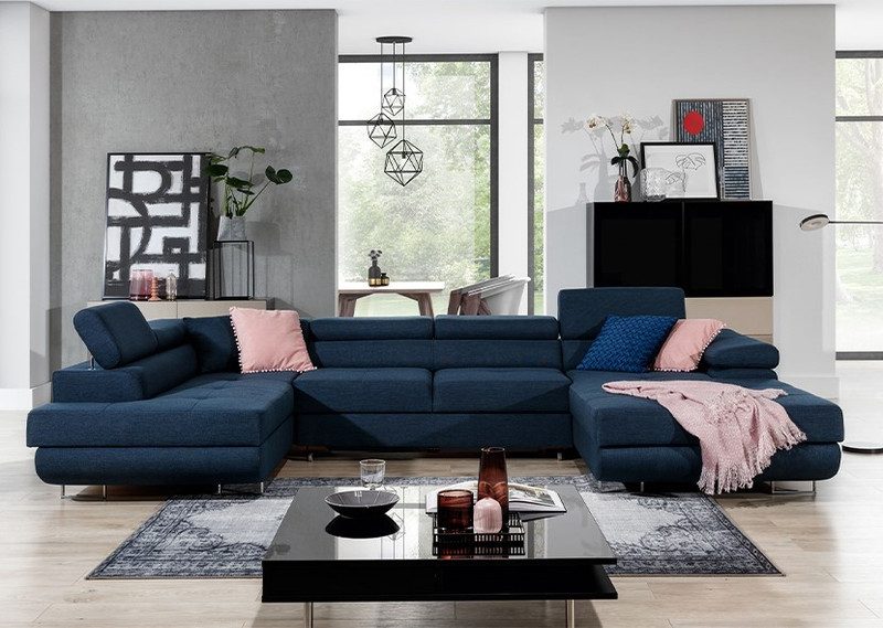 Navy blue Cairnwell U-shaped sofa bed with adjustable headrests, pink and blue scatter cushions and a pink throw, in a grey living room with a black coffee table