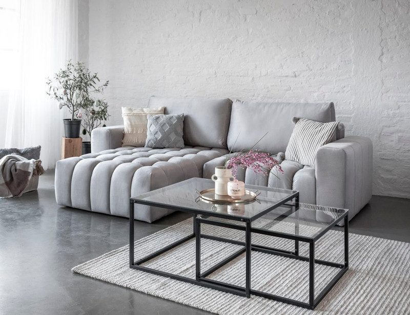 Light grey Ambleford corner sofa bed with a channel-tufted chaise on the left, cream, grey and striped cushions, glass nesting tables and a striped rug