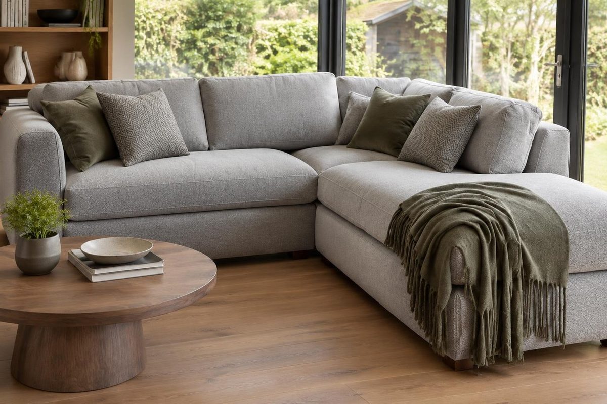 Grey Radstone corner sofa with a chaise end on the right, olive and grey textured cushions and a fringed olive throw, beside a round wooden pedestal coffee table and glass garden doors