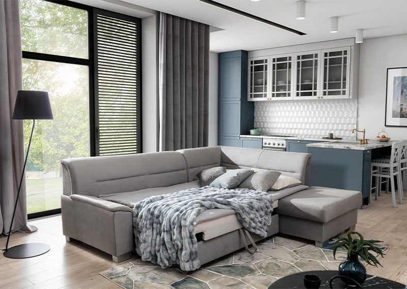 Light grey Mistral corner sofa bed opened out into a bed with pillows and a grey faux-fur throw, chaise on the right, in an open-plan living room with a kitchen behind