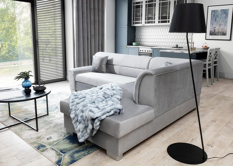 Light grey Mistral corner sofa bed with a grey faux-fur throw over the chaise, beside a black floor lamp and round coffee table in an open-plan living room