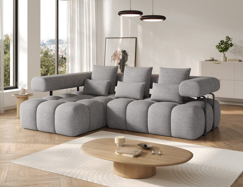Grey Ursaline corner sofa bed with the chaise on the left, three large back cushions and three smaller scatter cushions, beside an oval oak coffee table